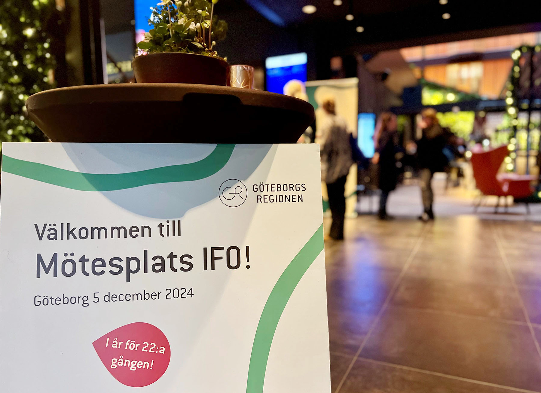 Skylt med texten Välkommen till mötesplats IFO. I bakgrunden syns deltagare