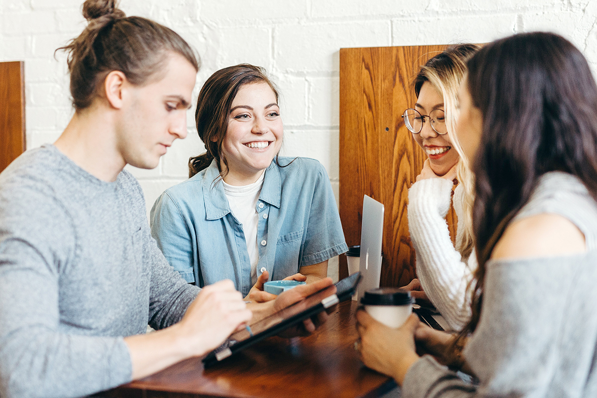 En grupp elever, tre tjejer och en kille, sitter vid ett bord. De dricker kaffe, har en dator och ipad framför sig, tittar på varandra och ler.