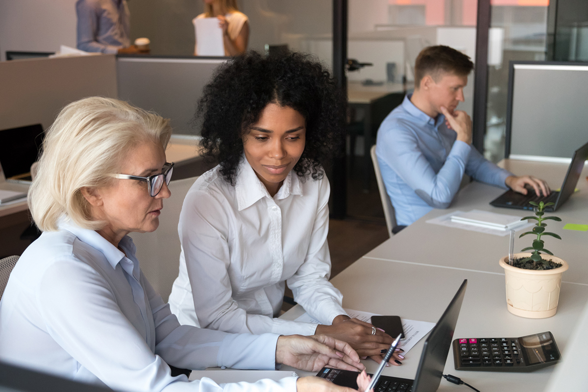 Två kvinnor sitter vid ett skrivbord i kontorsmiljö. De är klädda i blå och vit skjorta och tittar ner på en skärm tillsammans. Kvinnan till vänster visar den andra något på skärmen.