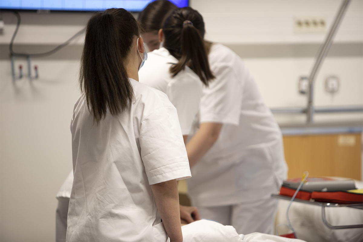 Undersköterskor arbetar meden patient. På bilden ser man bara deras ryggar. De har vita rockar på sig och tittar på en skärm. 