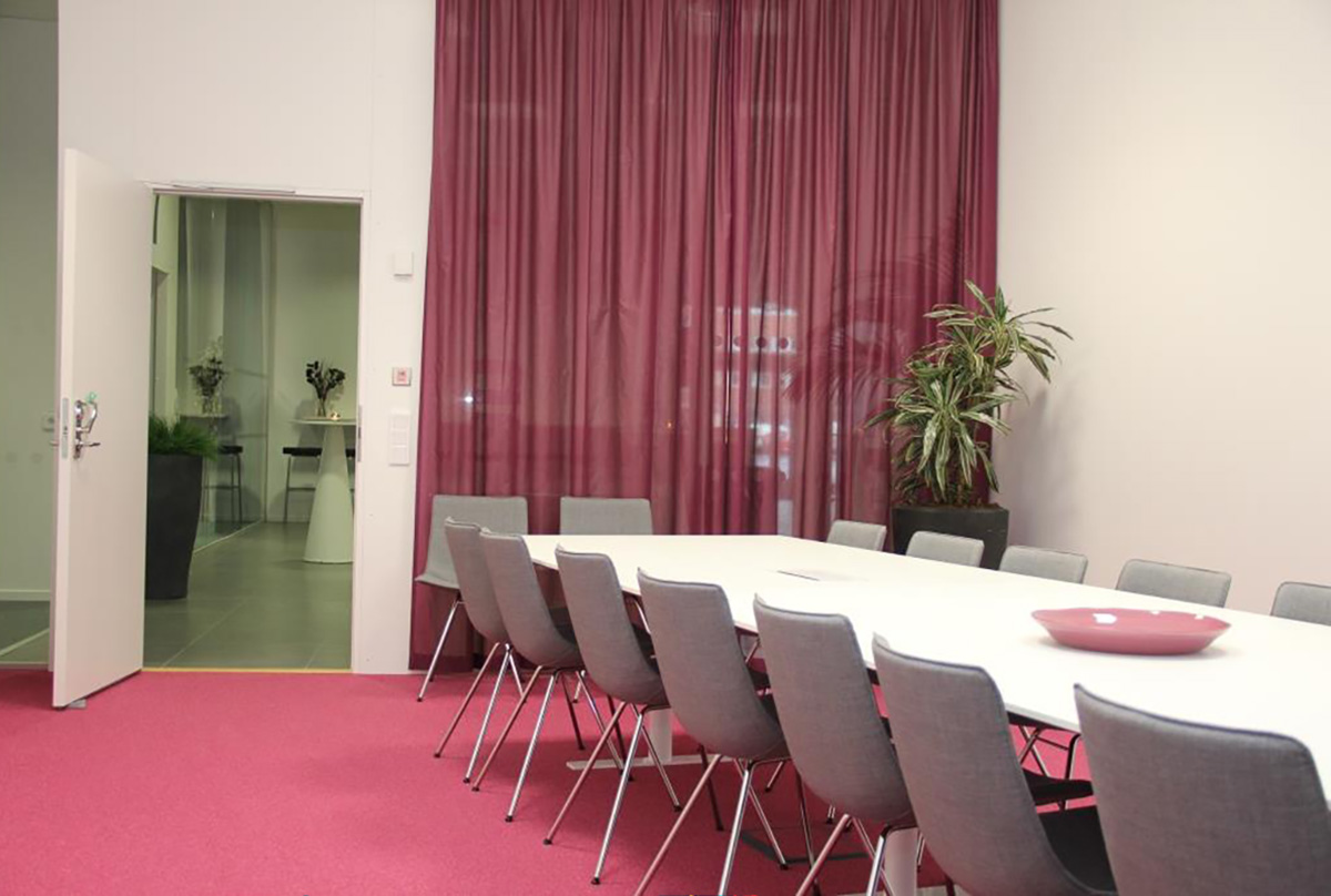 Interiör från ett av GR:s konferensrum. Rummet har en röd heltäckningsmatta och röda gardiner. Till höger står ett vitt konferensbord med grå stolar runt omkring.